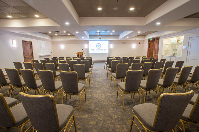 A wide meeting room with gray chairs, a projector screen, and a podium.