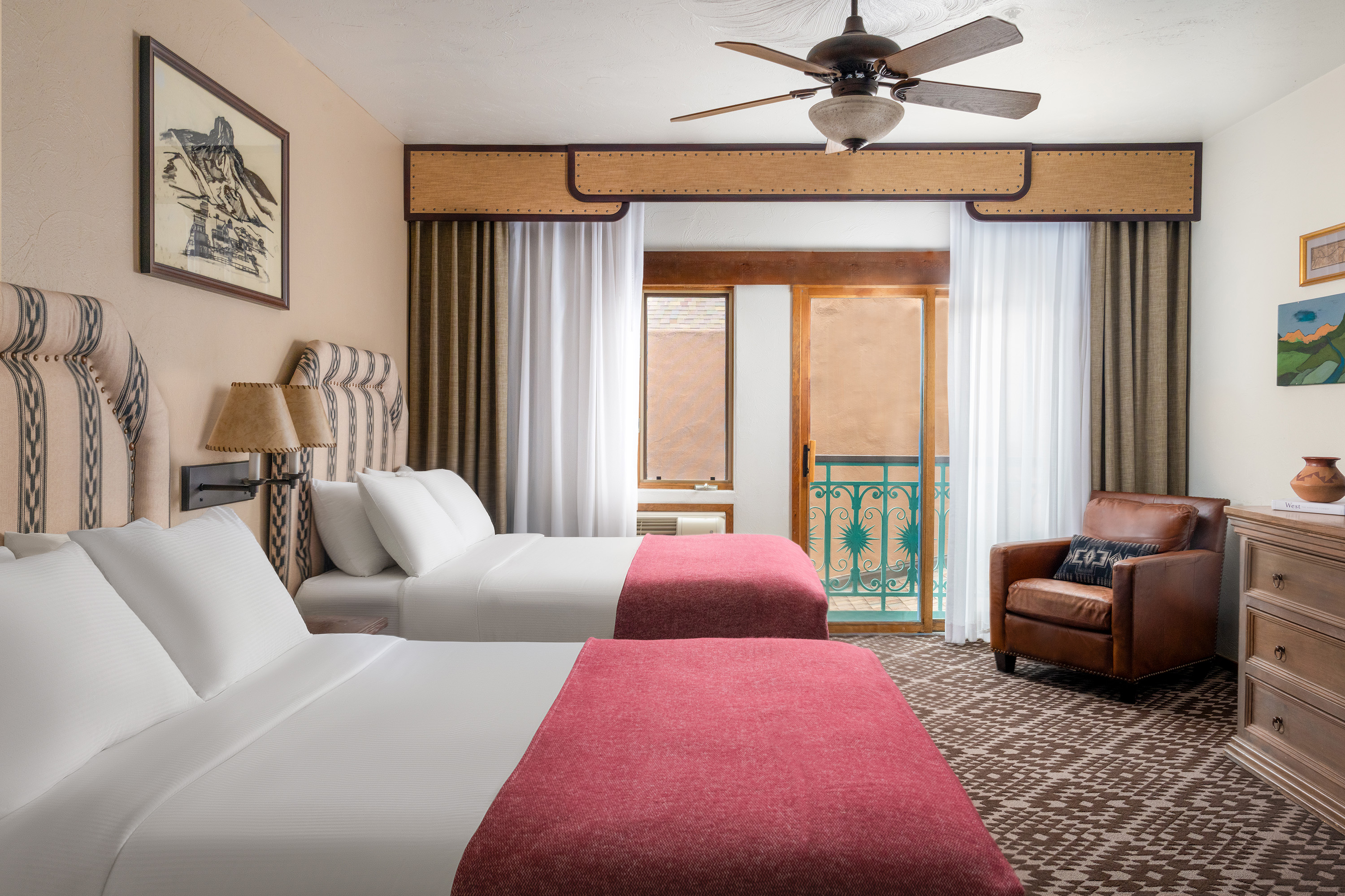 A hotel room with two double beds, a balcony door, a brown leather chair, and a ceiling fan.