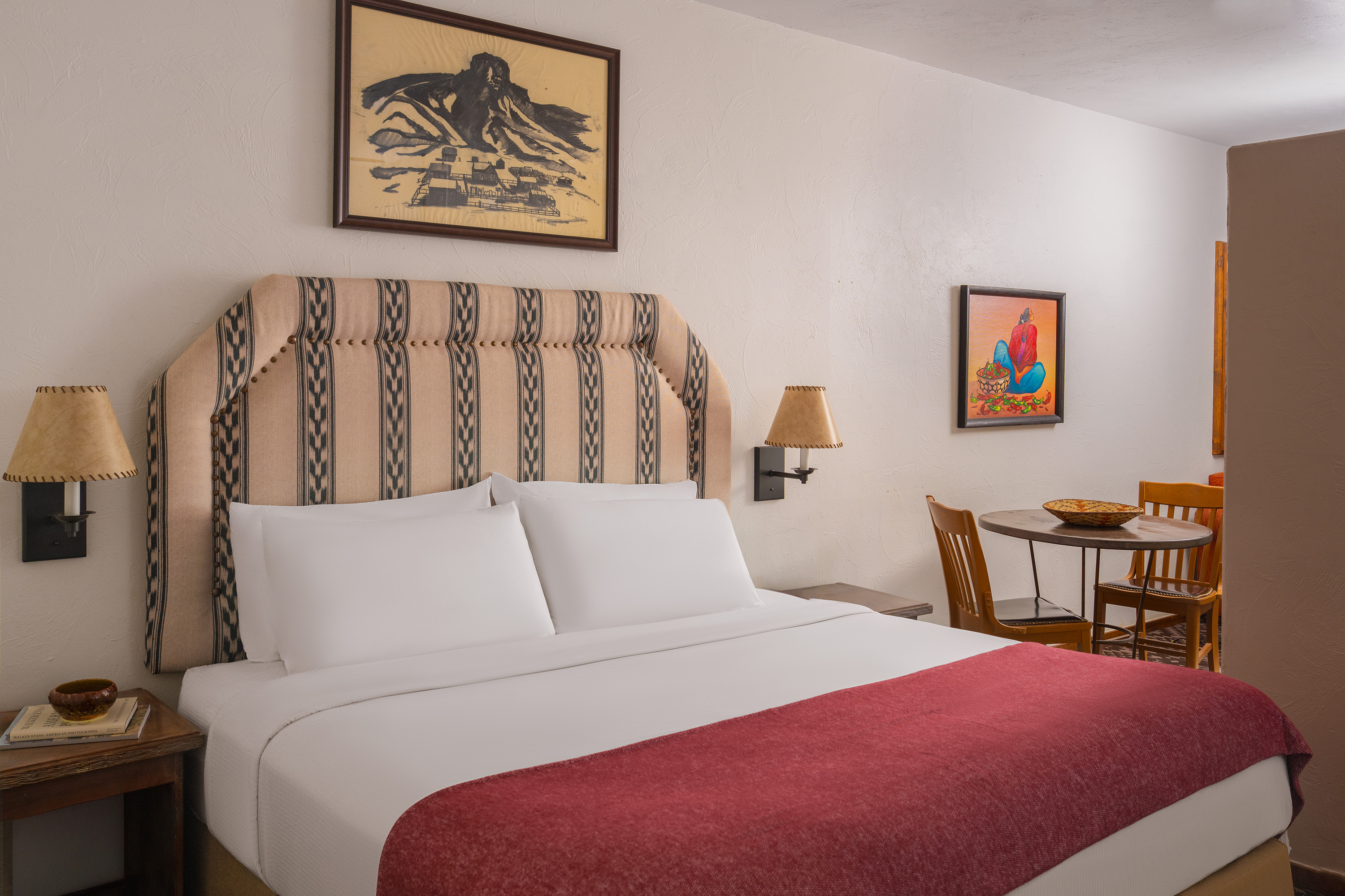 A hotel room with a bed, nightstand, table, chairs, and artwork on the walls.