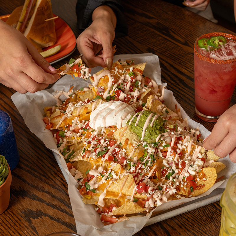 A platter of nachos with cheese, salsa, guacamole, and sour cream on a wooden table with hands reaching for it.